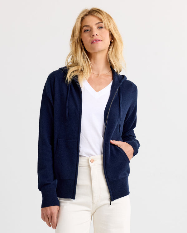 LIFESTYLE / EDITORIAL: Woman wearing navy zip-up cashmere hoodie over white V-neck shirt and cream pants against white