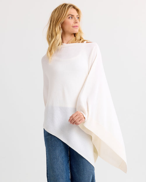 Woman wearing ivory asymmetrical cashmere poncho with blue jeans, standing against white background