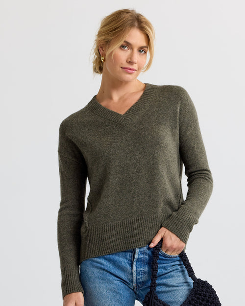 Woman wearing loden green V-neck cashmere sweater and blue jeans holding a black woven bag against white background