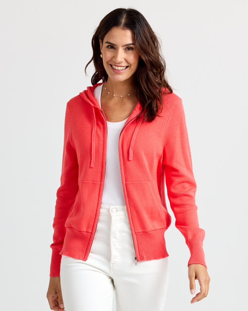 Front view image of a woman wearing a coral zip-up cashmere hoodie and white pants smiling against a white background