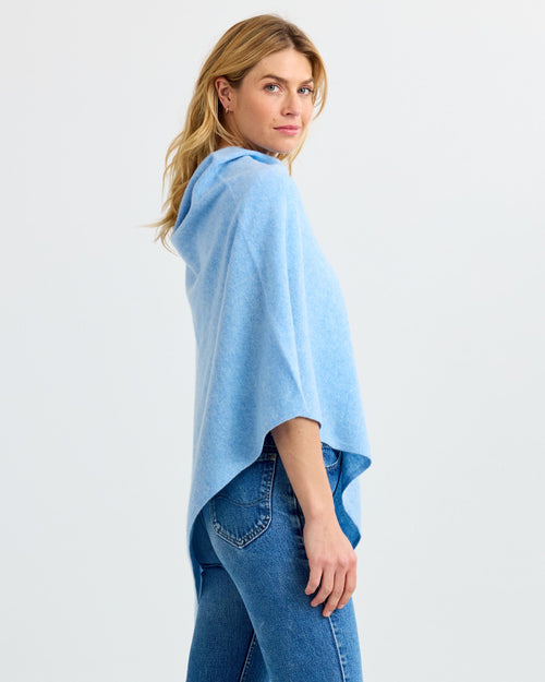 LIFESTYLE / EDITORIAL: Woman wearing light blue cashmere poncho with hood and blue jeans, side view against white background