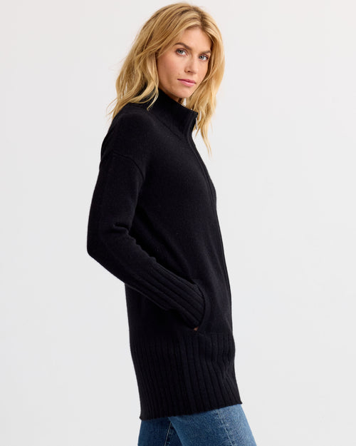 LIFESTYLE / EDITORIAL: Woman in black long cashmere sweater with pockets, standing sideways against white background