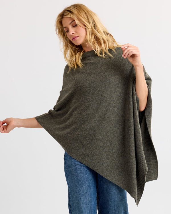 Side view image of a woman wearing dark olive asymmetrical cashmere poncho with blue jeans against white background