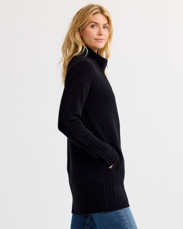 LIFESTYLE / EDITORIAL: Woman in black long cashmere sweater with high collar, side view, hands in pockets, white background