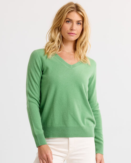 LIFESTYLE / EDITORIAL: Woman wearing a green V-neck cashmere sweater and white pants against a plain background