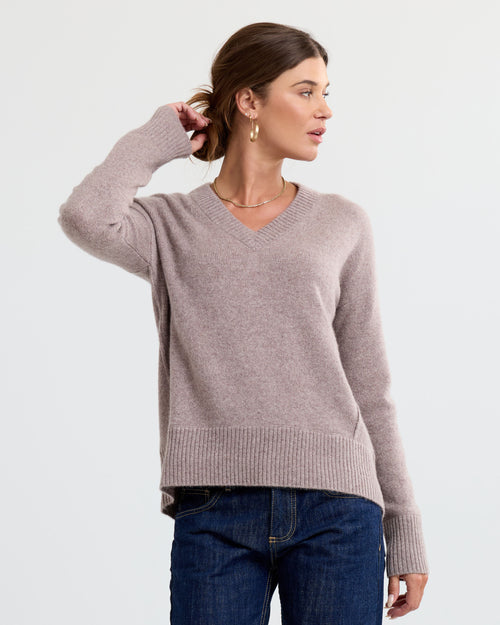 LIFESTYLE / EDITORIAL: Woman wearing a latte-colored V-neck cashmere sweater and dark jeans, posing against a plain