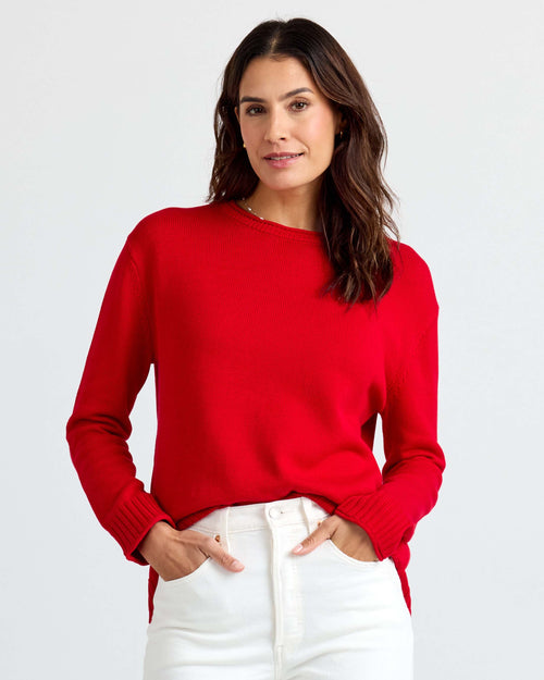 LIFESTYLE / EDITORIAL: Woman wearing red cashmere sweater and white pants, standing with hands in pockets against white