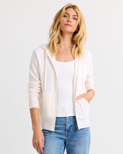 LIFESTYLE / EDITORIAL: Woman wearing ivory zip-up cashmere hoodie, white tank top, and blue jeans against white background