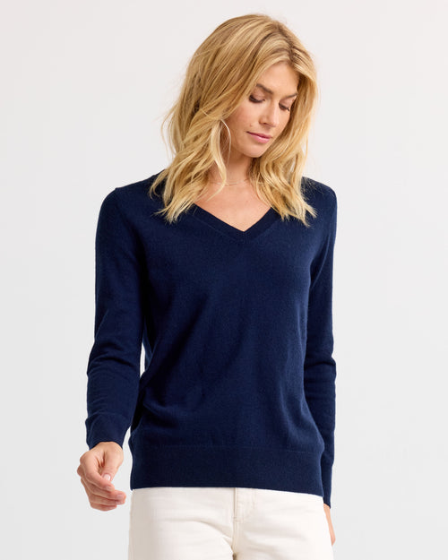 LIFESTYLE / EDITORIAL: Woman wearing navy V-neck cashmere sweater and white pants, standing against plain background