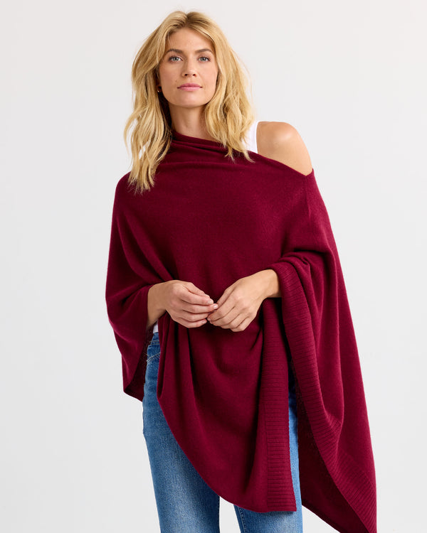 LIFESTYLE / EDITORIAL: Woman wearing off-shoulder burgundy cashmere poncho with blue jeans against white background