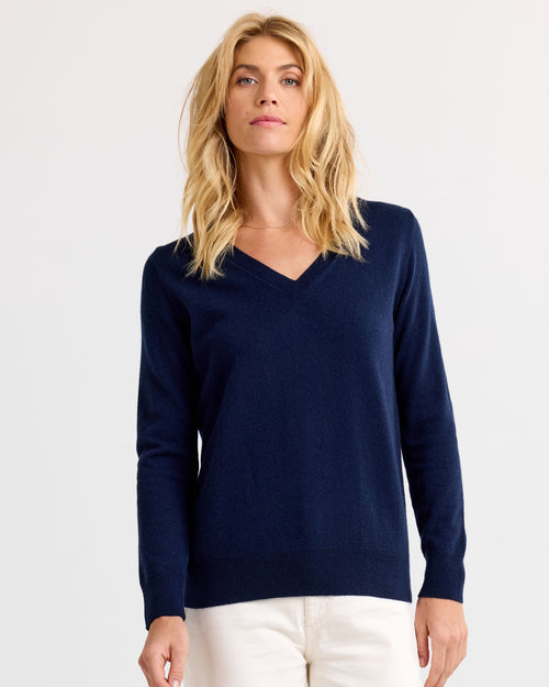 Front view image of a woman wearing a navy V-neck cashmere sweater with white pants against a white backdrop