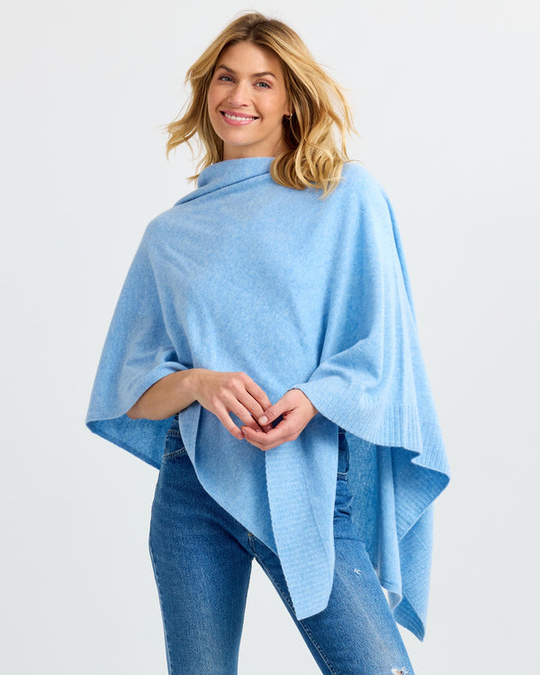 LIFESTYLE / EDITORIAL: Woman wearing light blue cashmere poncho and jeans, smiling against plain background