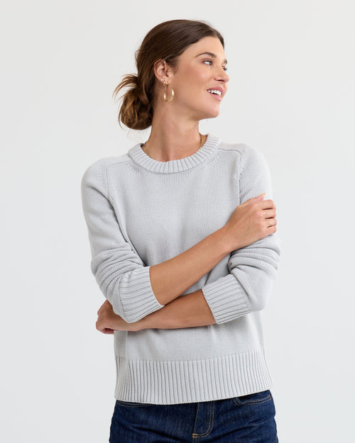 Woman wearing light grey cashmere sweater with ribbed cuffs and hem, paired with dark jeans, looking to side
