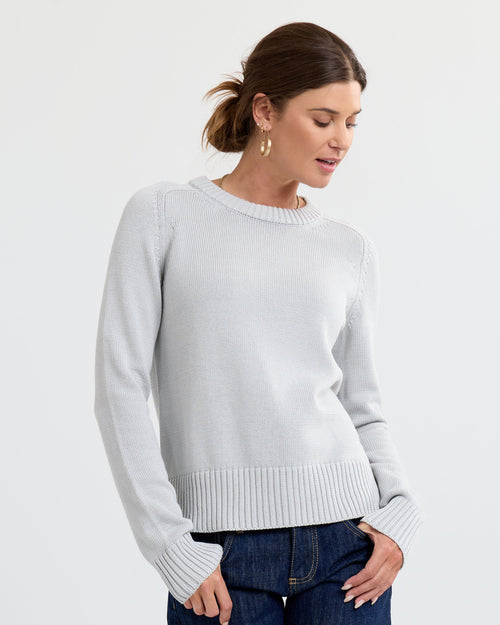 LIFESTYLE / EDITORIAL: Woman wearing light grey cashmere sweater and blue jeans, standing against white background