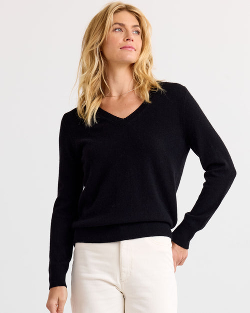 Three-quarter view image of a woman wearing a black V-neck cashmere sweater and white pants, looking slightly up.