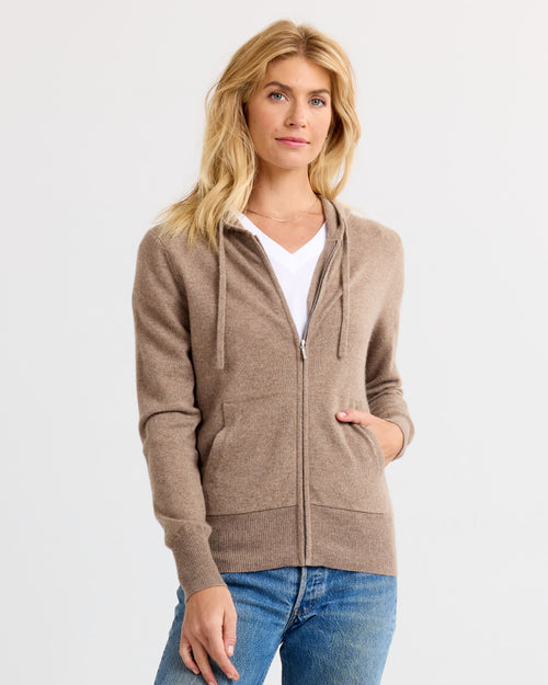 Woman wearing driftwood-colored zip-up cashmere hoodie over white tee and blue jeans against white background