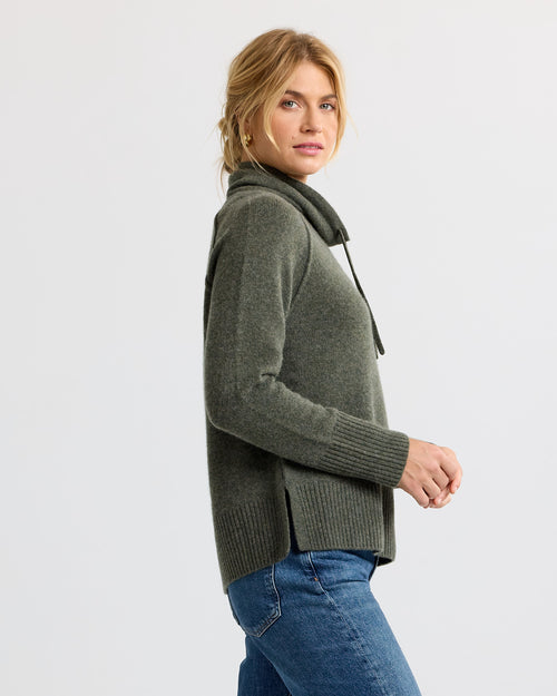 Woman wearing dark olive cashmere sweater with cowl neck and side slits, paired with blue jeans, side profile view