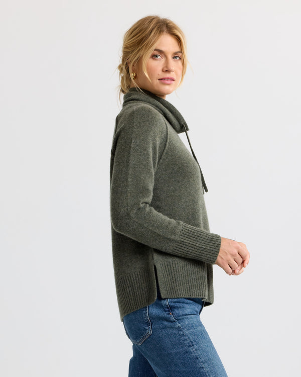 Woman wearing dark olive cashmere sweater with cowl neck and side slits, paired with blue jeans, side profile view
