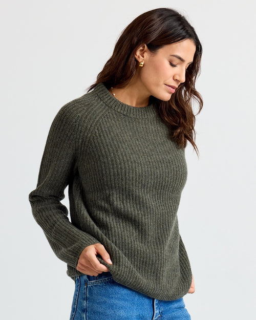 LIFESTYLE / EDITORIAL: Woman wearing dark olive ribbed cashmere sweater and blue jeans, looking down with hands near pockets