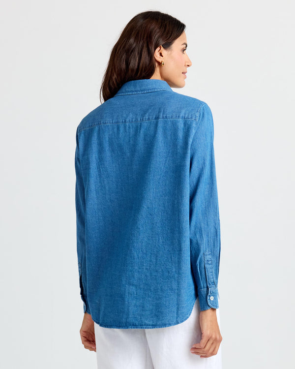 Woman wearing medium wash blue denim shirt, shown from the back, standing against a plain light background