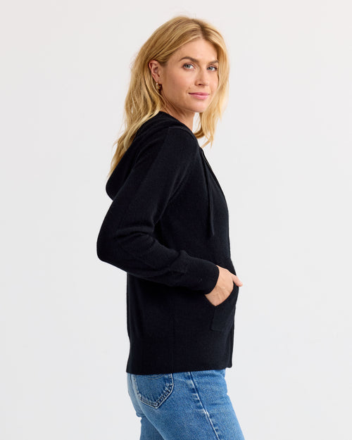 LIFESTYLE / EDITORIAL: Woman in black cashmere hoodie and blue jeans, side view, against plain white background