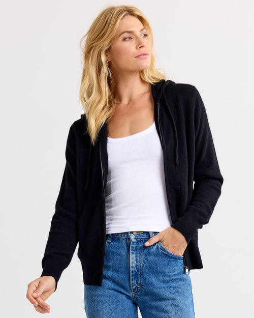 Three-quarter view image of a woman wearing black zip-up cashmere hoodie over white tank and blue jeans
