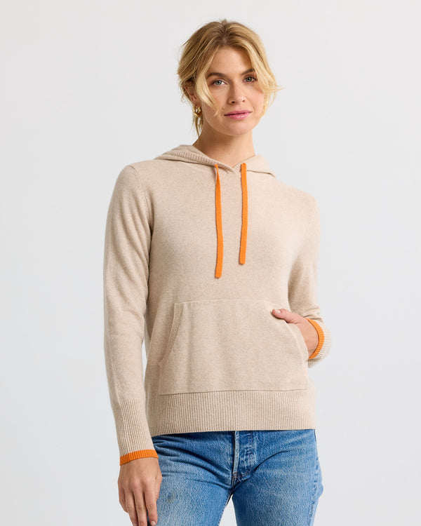 LIFESTYLE / EDITORIAL: Woman wearing sand-colored cashmere hoodie with orange drawstrings and cuffs, paired with blue jeans