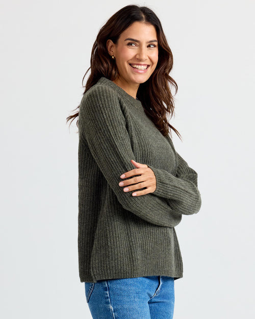 LIFESTYLE / EDITORIAL: Woman smiling, wearing dark olive ribbed cashmere sweater and blue jeans against white background