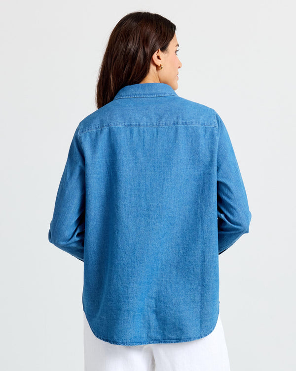 LIFESTYLE / EDITORIAL: Woman wearing medium wash blue denim shirt, back view, white pants, standing against white background