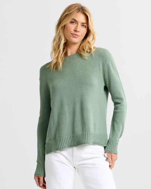 Woman wearing sage green cashmere sweater and white pants against plain light background