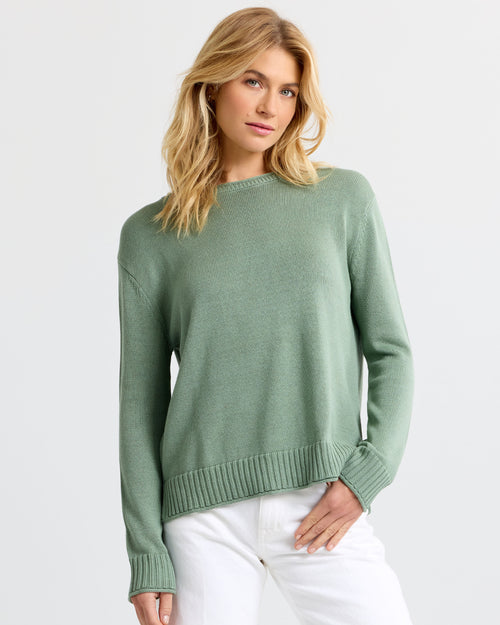 Front view image of a woman wearing a sage green cashmere sweater and white pants standing against a light background