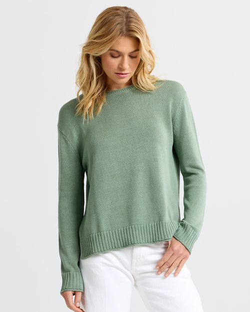 Front view image of a woman wearing a sage green cashmere sweater and white pants, looking down against a plain background