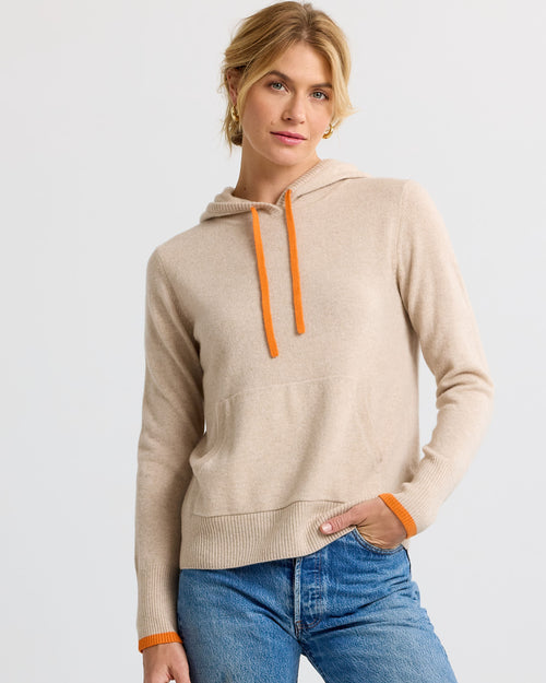 LIFESTYLE / EDITORIAL: Woman wearing beige cashmere hoodie with orange drawstrings and cuffs, paired with blue jeans