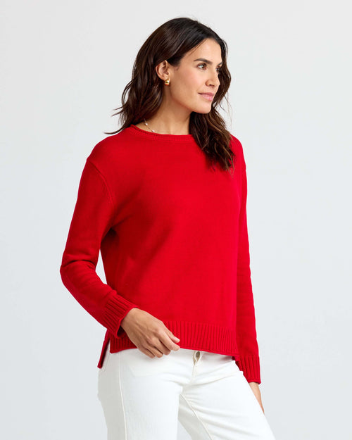 LIFESTYLE / EDITORIAL: Woman wearing red cashmere sweater and white pants, standing against a plain light background