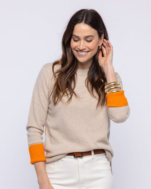 LIFESTYLE / EDITORIAL: Woman smiling, wearing beige cashmere sweater with orange cuffs, white pants, and gold bangles