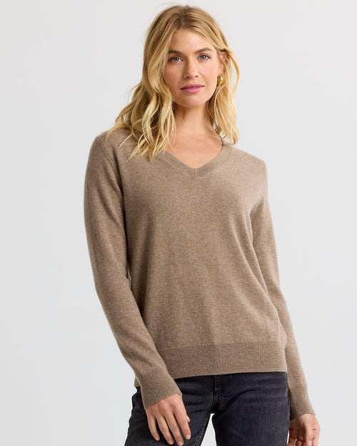 Three-quarter image of a woman wearing a driftwood-colored V-neck cashmere sweater and dark jeans against plain background