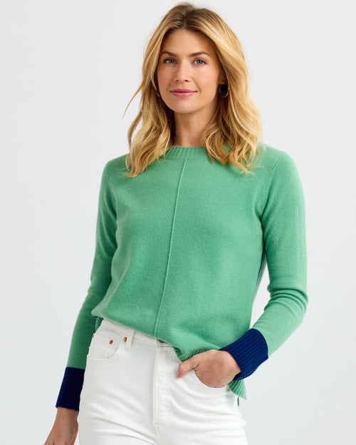 LIFESTYLE / EDITORIAL: Woman wearing green cashmere sweater with navy cuffs and white pants, standing against white