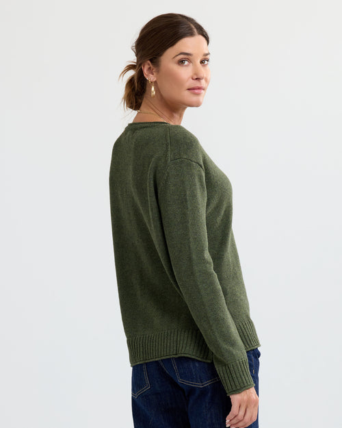 Woman wearing olive green cashmere sweater, turned slightly, with dark blue jeans, plain background