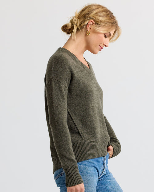 Woman wearing olive green V-neck cashmere sweater and blue jeans, side profile against plain background