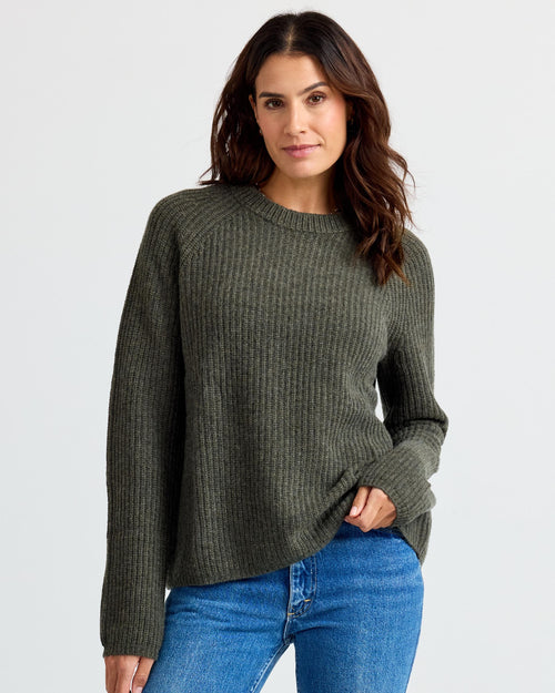 Front view image of a woman wearing a dark olive ribbed cashmere sweater and blue jeans against white background