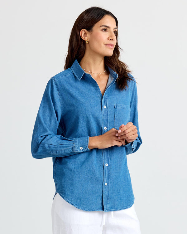Three-quarter view image of a woman wearing a medium wash denim shirt, hands clasped, looking sideways.