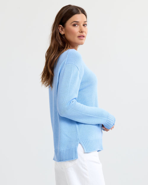 LIFESTYLE / EDITORIAL: Woman in light blue cashmere sweater and white pants, posing sideways against a plain background
