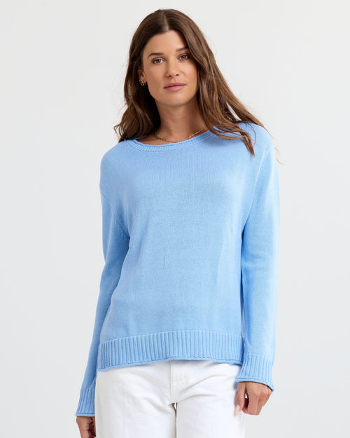Front-facing image of a woman wearing a light blue cashmere sweater and white pants against a plain background