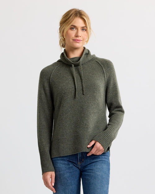 Woman wearing dark olive cashmere cowl neck sweater with drawstrings and blue jeans, standing against white background