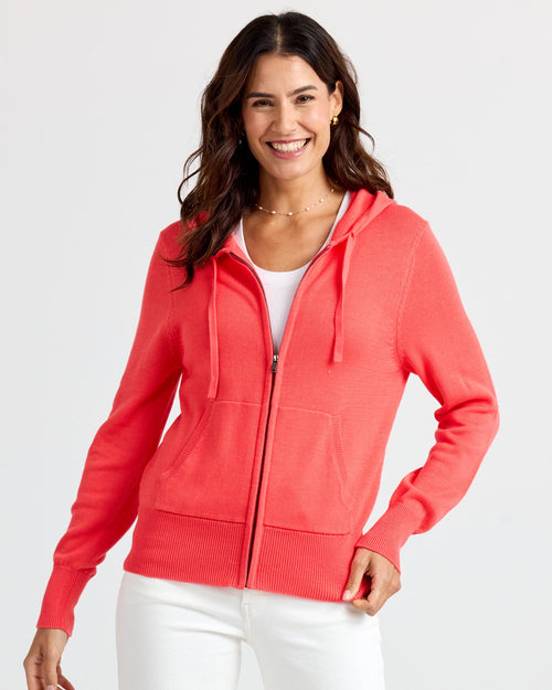 LIFESTYLE / EDITORIAL: Smiling woman wearing coral zip-up cashmere hoodie and white pants against plain background