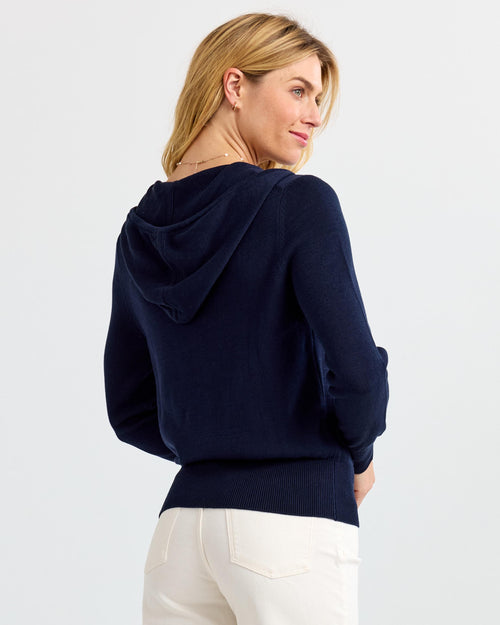Rear three-quarter view image of a woman wearing a navy cashmere hoodie with white pants against plain background