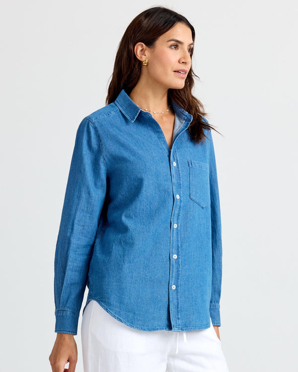 LIFESTYLE / EDITORIAL: Woman wearing medium wash denim shirt and white pants, standing against a plain background