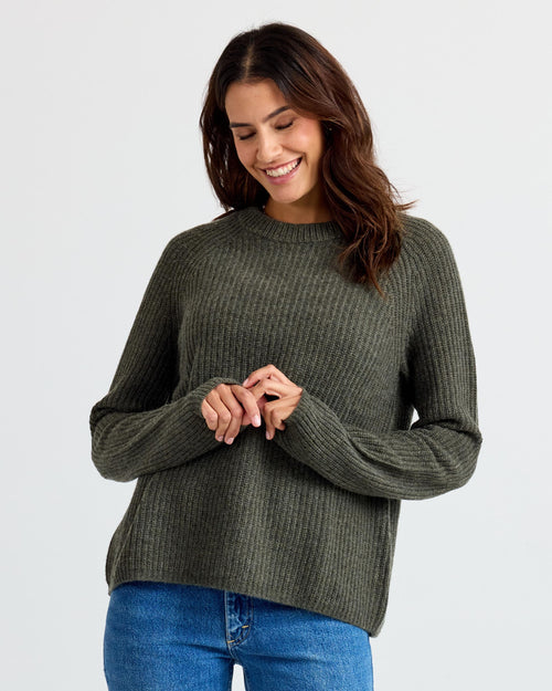 LIFESTYLE / EDITORIAL: Woman wearing dark olive ribbed cashmere sweater and blue jeans, smiling with hands clasped