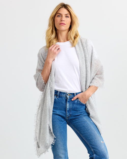 Three-quarter view image of a woman wearing a light gray cashmere shawl over a white tee and blue jeans.