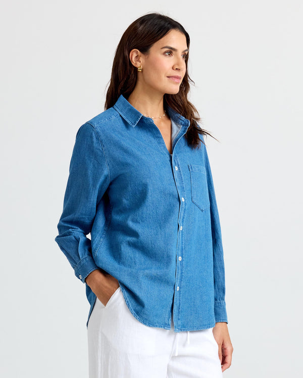 Three-quarter view image of a woman wearing a medium wash denim shirt and white pants, standing with hands in pockets